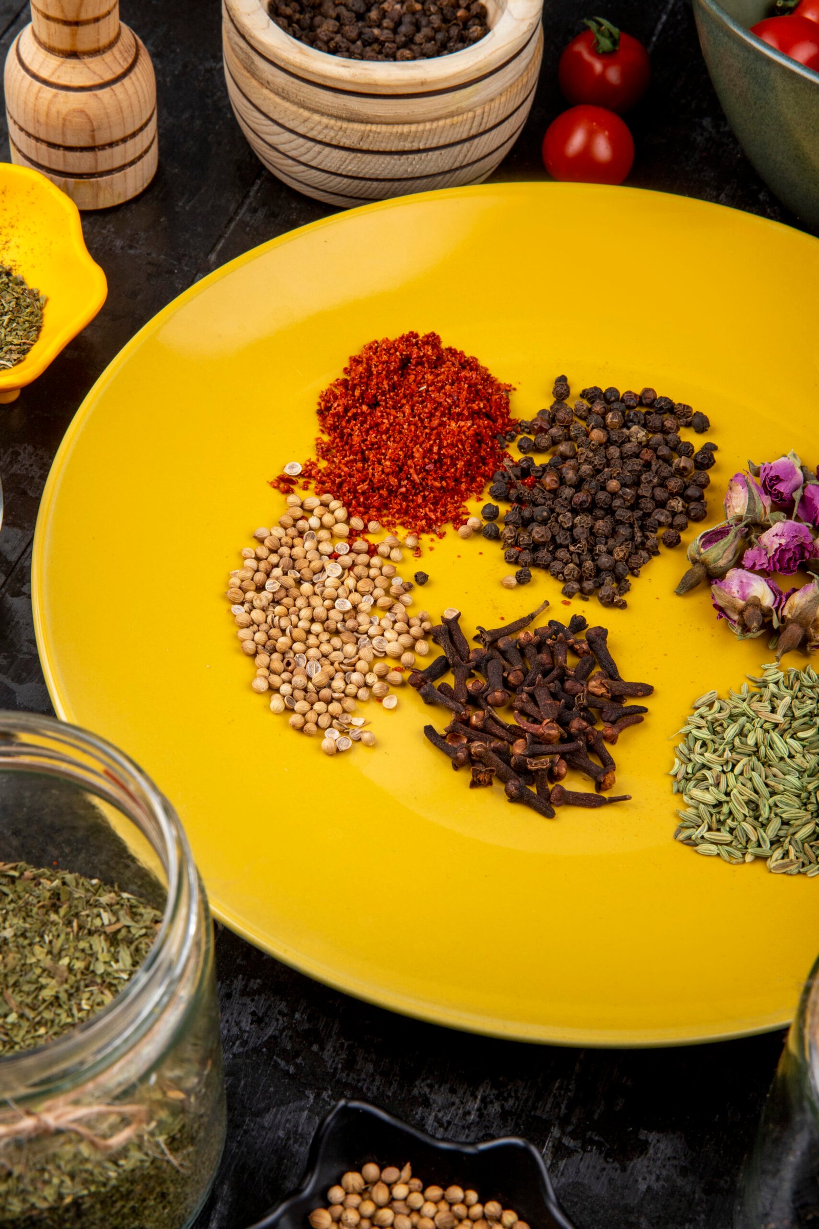 side view of a set of spices and herbs peppercorns anise seeds clove red chili pepper flakes and tea rose buds on yellow plate
